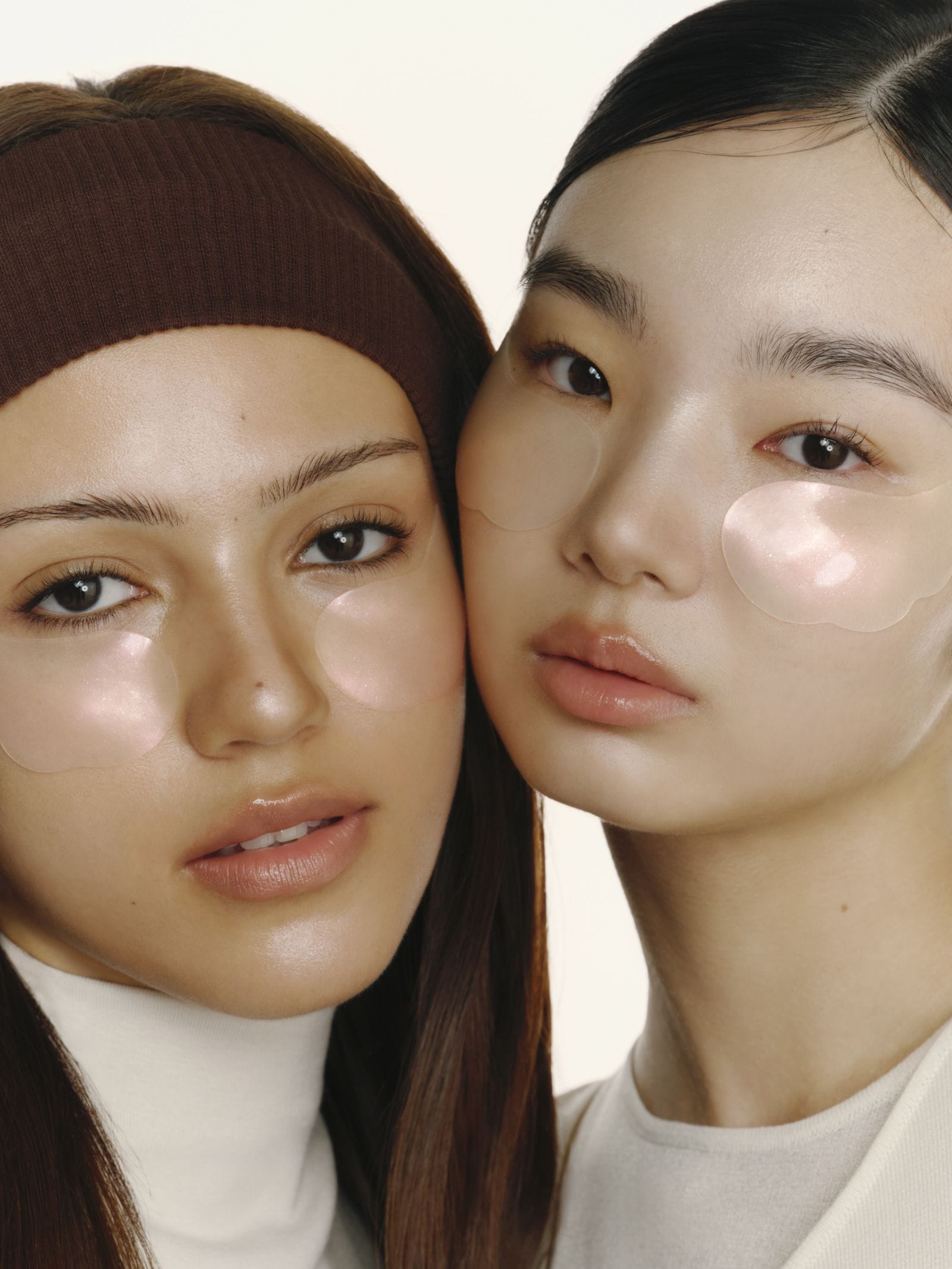 Two women in a close-up portrait wearing clear under-eye patch masks and a brown ribbed headband, set against a clean off-white background.